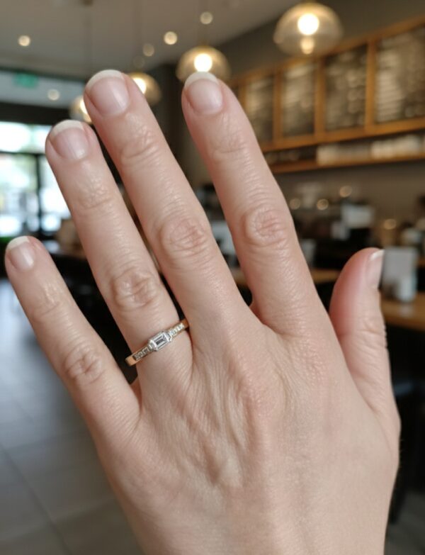 Kädessä siro 18K kultainen timanttisormus, jossa suorakulmainen keskikivi.